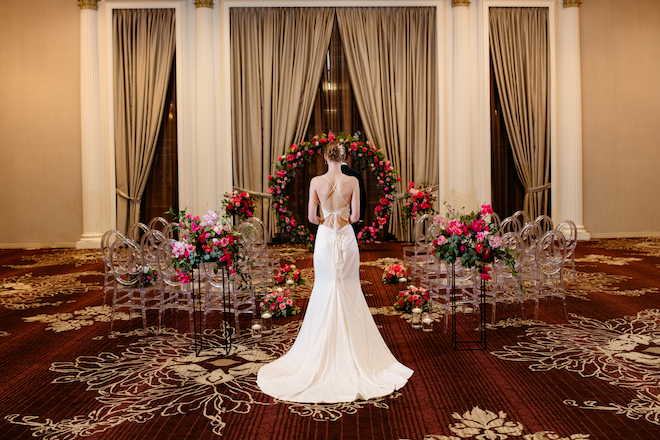 The bride facing the ceremony space covered in greenery and pink florals with ghost chairs. 