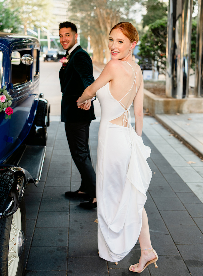 The bride looking back smiling while holding the groom's hand in front of a vintage blue car. 