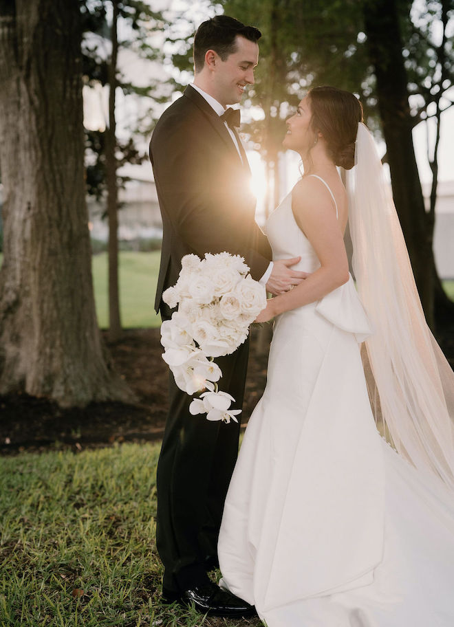 The bride and groom smiling at each other with the bride holding a bouquet of white florals. 