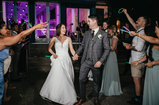 The bride and groom exit holding hands as they leave their wedding reception with a glow stick send-off. 