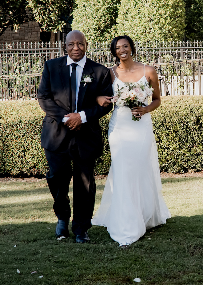 The bride is escorted down the aisle by her father at her outdoor wedding ceremony. 