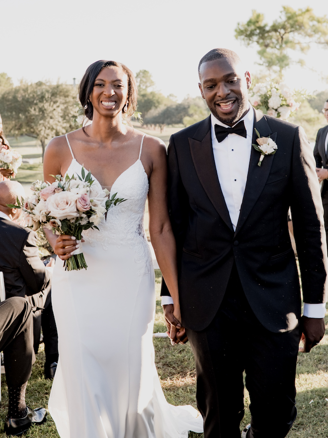 The bride and groom hold hands and smile as the walk down the aisle as newlyweds. 