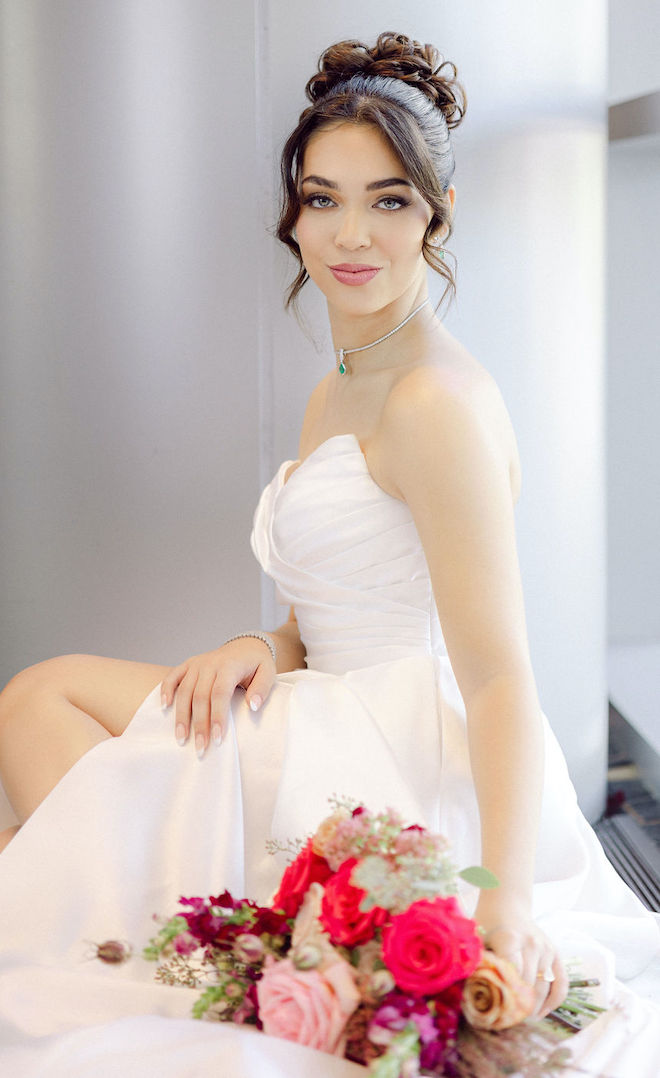 The bride sits in front of a window as she holds her bouquet and soft smiles at the camera. 