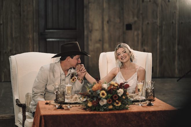 The groom kisses the brides hand while they sit at their table in the reception hall. 