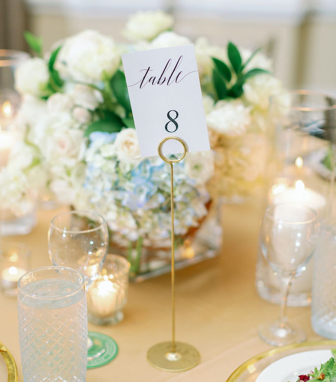 A centerpiece of white and blue florals with a "Table 8" sign in front of it with gold accents. 