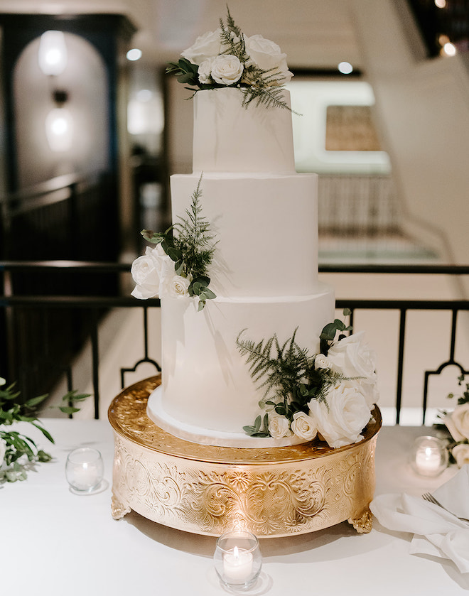 A three-tier white wedding cake decorated in white roses and greenery is surrounded by candlelight at the bride and groom's wedding reception.