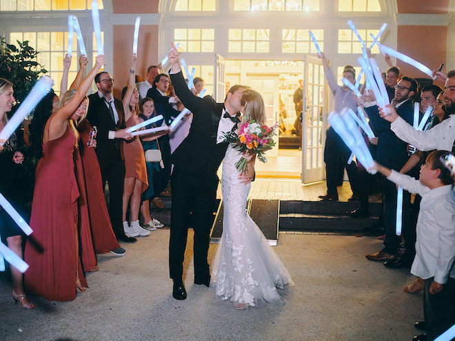 The bride and groom kissing as the guests wave glow sticks on either side of them. 