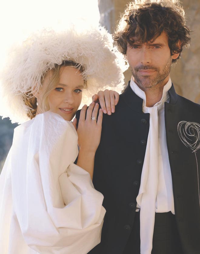 A bride wearing a white fluffy hat with her hands resting on the grooms shoulder. 