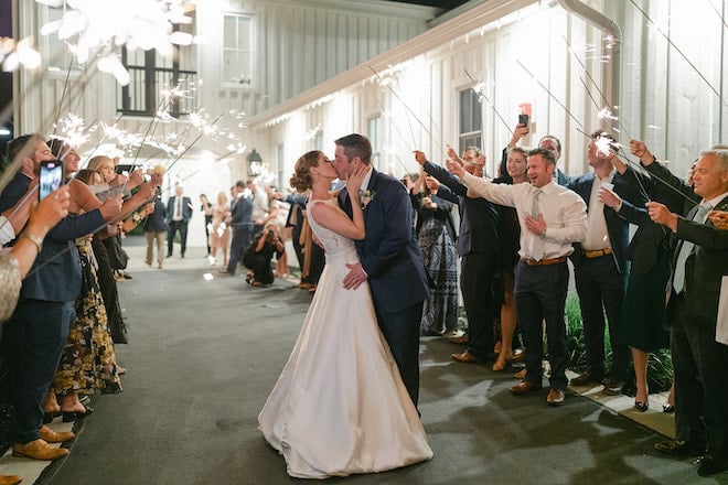 The bride and groom kissing while guests hold sparklers for their sendoff. 