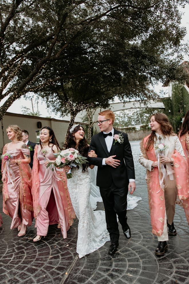 The bride and groom lock arms as they walk outside the wedding venue with the bridal party wearing traditional Bengali attire. 