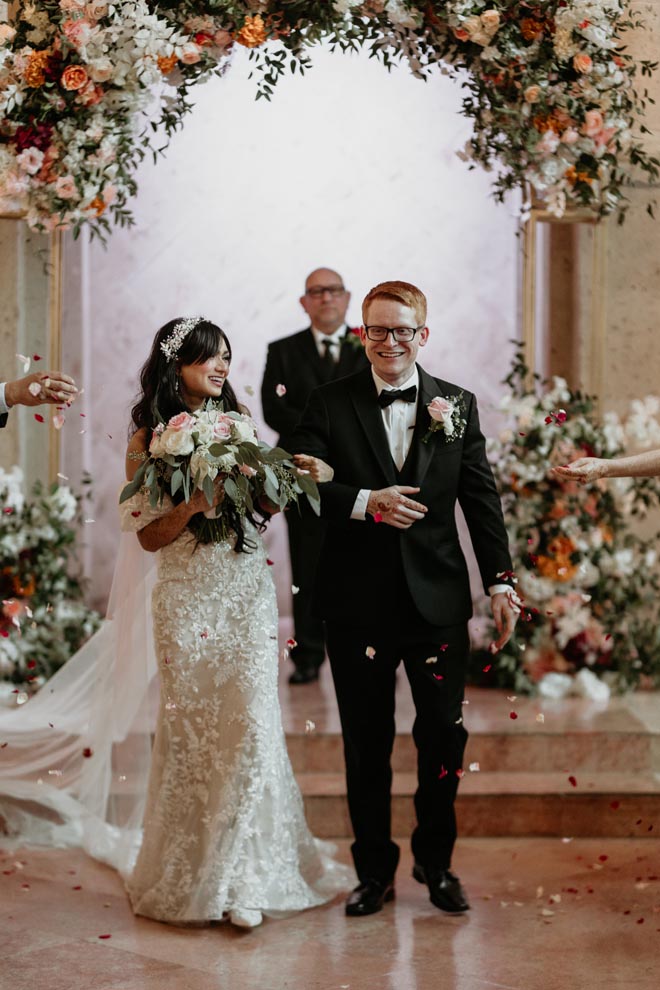 The bride and groom lock arms as they walk down the aisle as their guests throw flower petals in the air. 