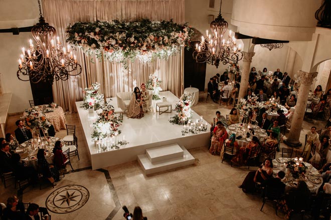 The bride and groom sit on the stage at their wedding reception as wedding guests watch. 