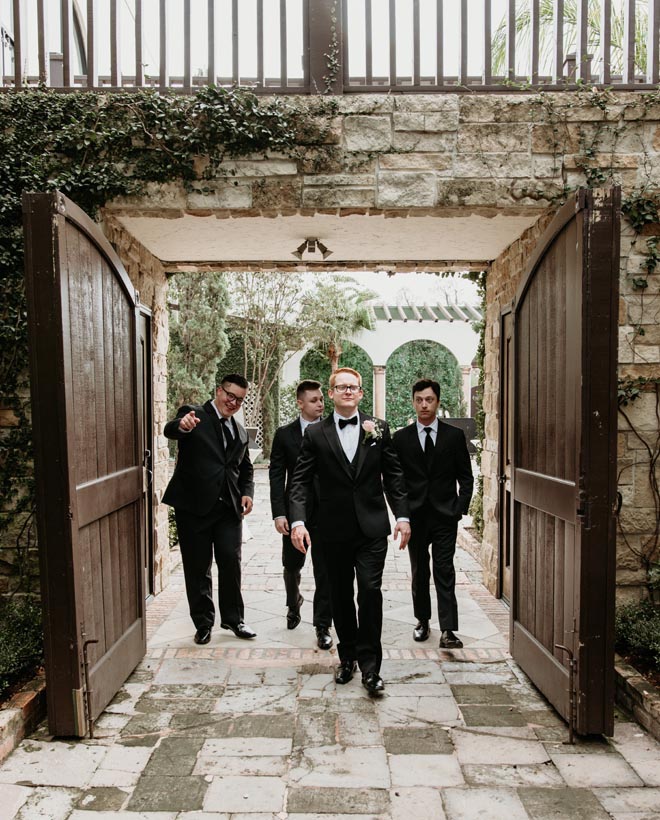 The groom and his groomsmen wearing tuxedos walk outside the wedding venue in Houston, The Bell Tower on 34th.