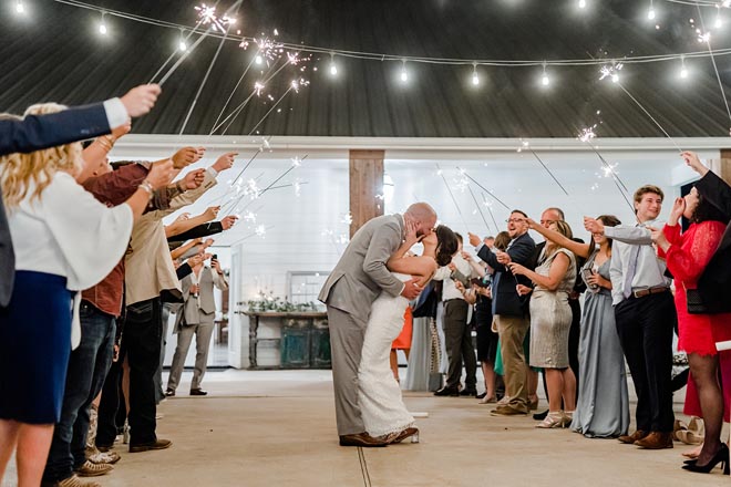 Wedding guests wave sparklers in the air as the bride and kiss exiting their countryside wedding reception. 