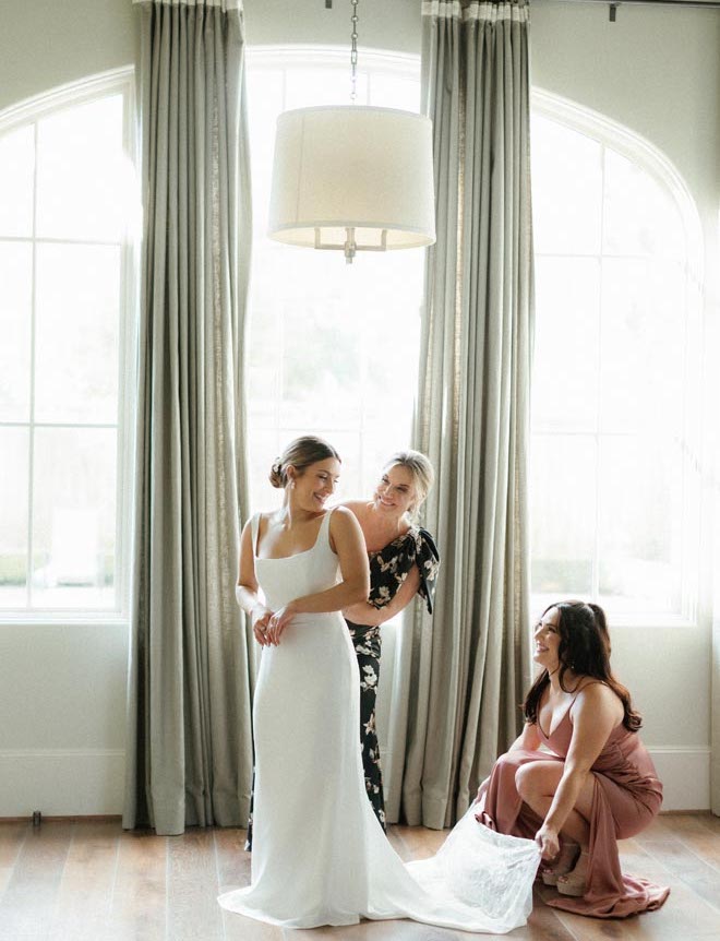 The bride's mother and sister help the bride zip into her wedding gown.