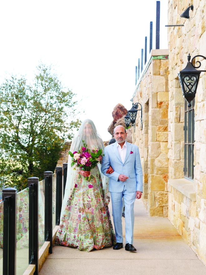 The bride walks down the aisle with her father as she wears a veil over her head. 