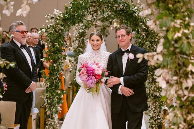 The bride and her father walking down the aisle under greenery archways. 