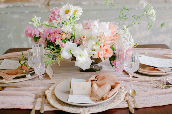 A table scape with a pink and white bouquet, beige plates and silver flatware with peach-colored linens. 