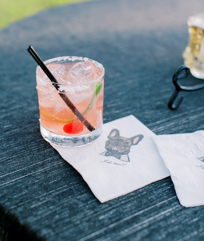 A pink cocktail sitting on a napkin wit a print of a french bulldog wearing a bowtie reading "I Do Too!!"