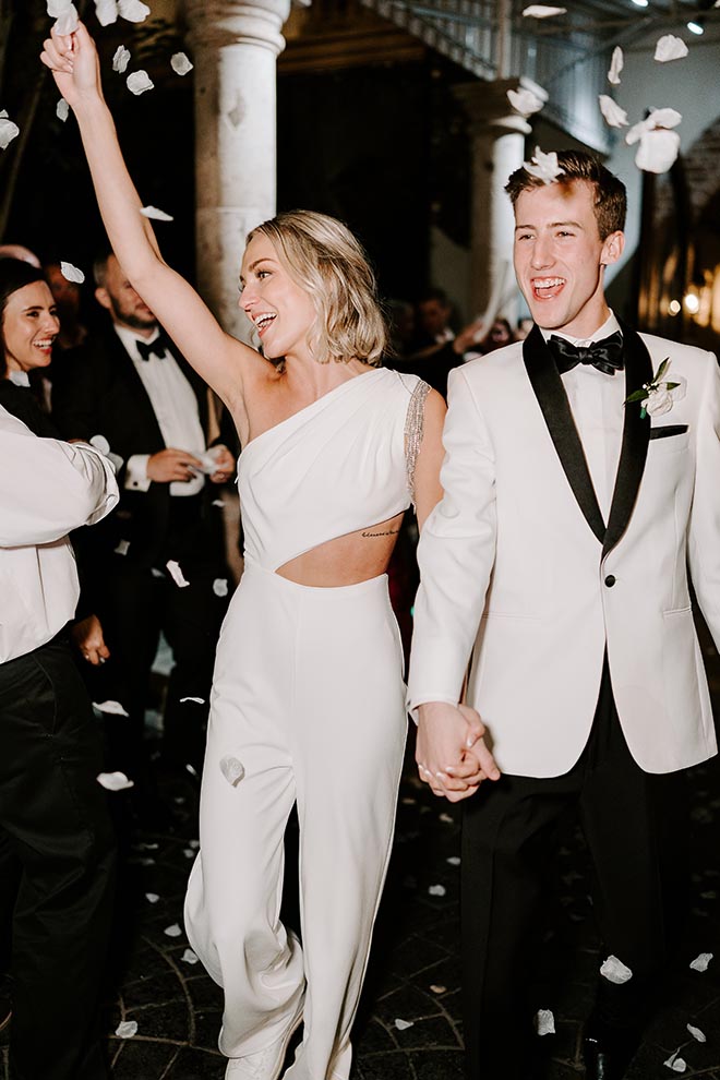 The bride wearing a white jumpsuit and holding the groom's hand while guests throw flower petals at them. 