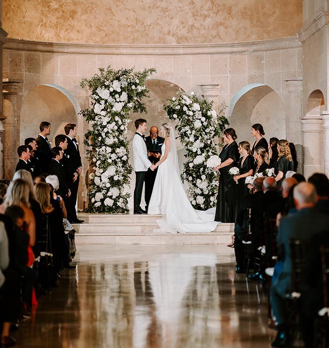 The bride and groom holding hands at the altar decorated with greenery and white floral installations. 