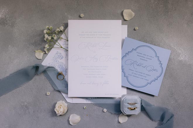 White and light blue wedding stationery are surrounded by white flower petals.