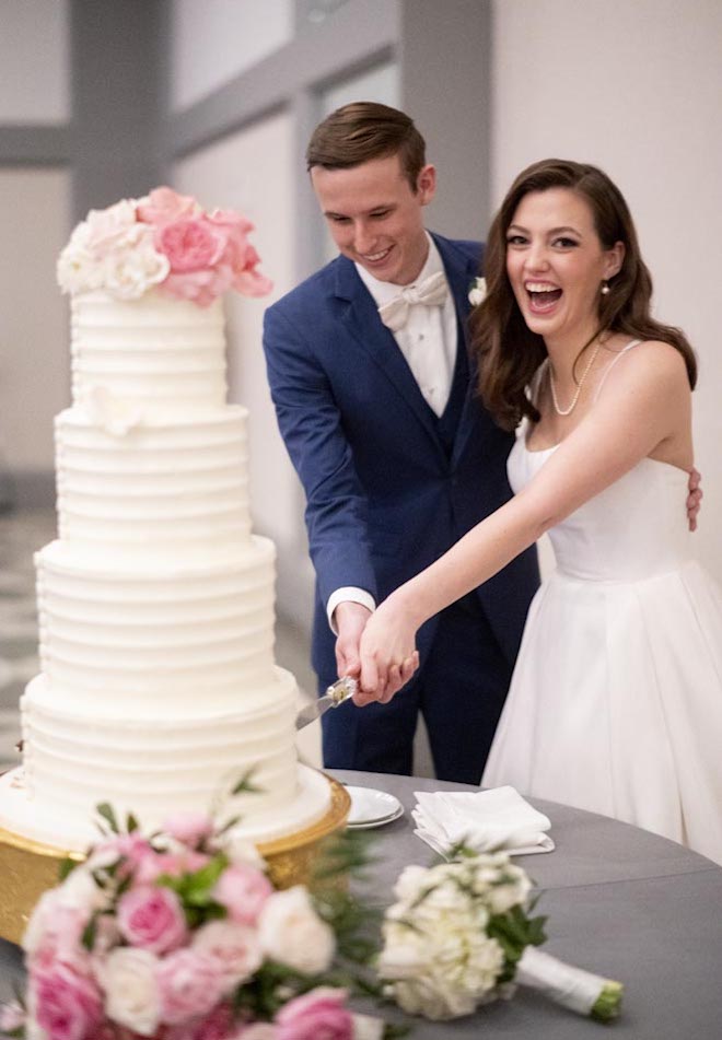 The bride and groom slice into their four-tier white wedding cake topped with pink roses. 