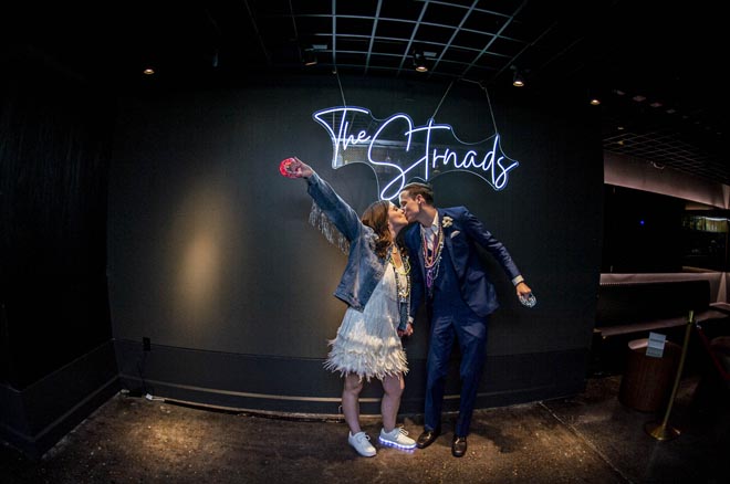 The bride and groom stand under a neon sign that says the couples last name "Strnad."