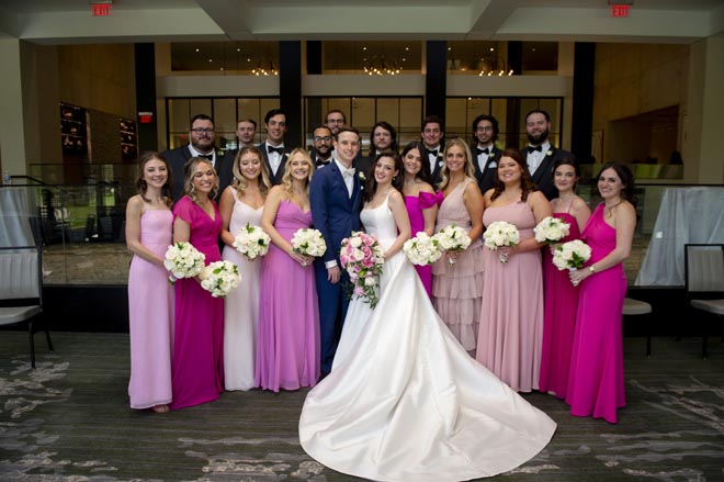 The wedding party smile next to the bride and groom wearing pink and navy attire. 