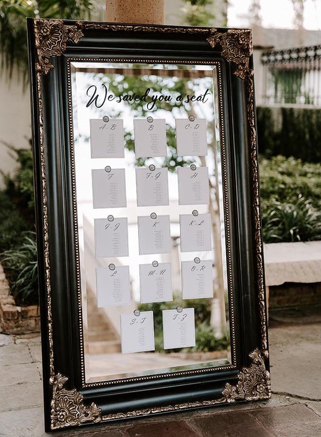 A black and gold mirror with writing that reads "We saved you a seat" with a seating chart attached to the mirror. 