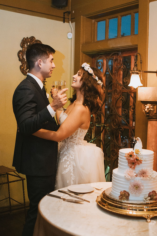 The bride and groom celebrate with a glass of champagne beside their wedding cake.