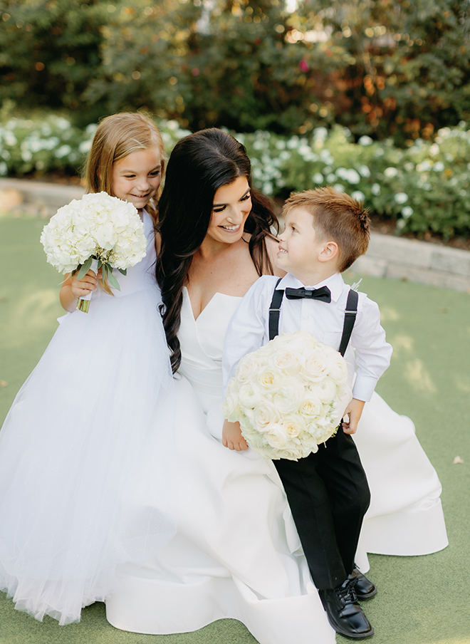 The bride smiling at two young children holding white bouquets. 