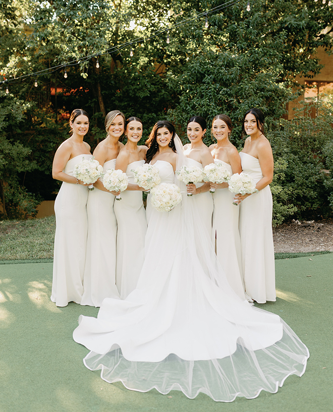 The bride smiling with her six bridesmaids, all holding white bouquets and wearing strapless white gowns. 