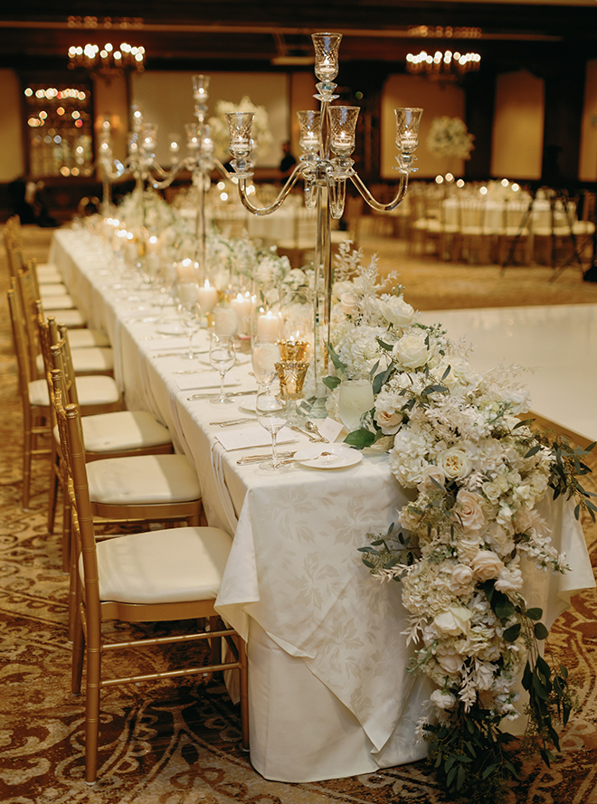 A long table with white linens, glass candle holders and a white floral arrangement in the ballroom of The Houstonian Hotel, Club & Spa for a traditional wedding. 