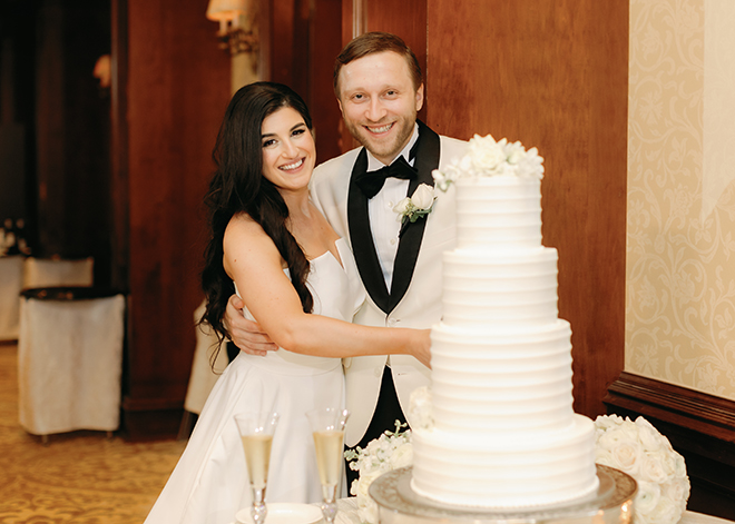 The bride and groom smiling next to a 4-tier white wedding cake by Susie's Cakes. 