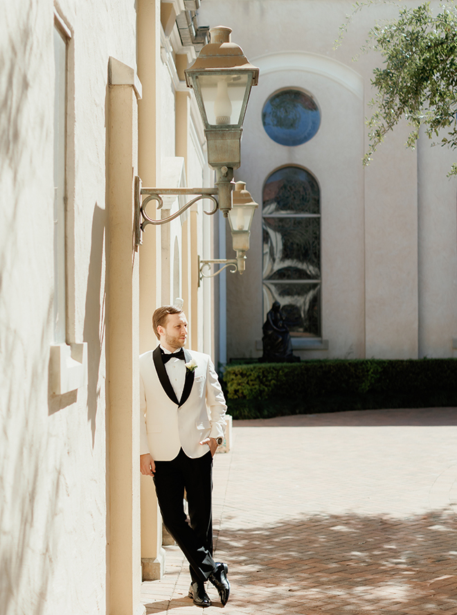 The groom wearing a white suit jacket leaning against the church wall. 