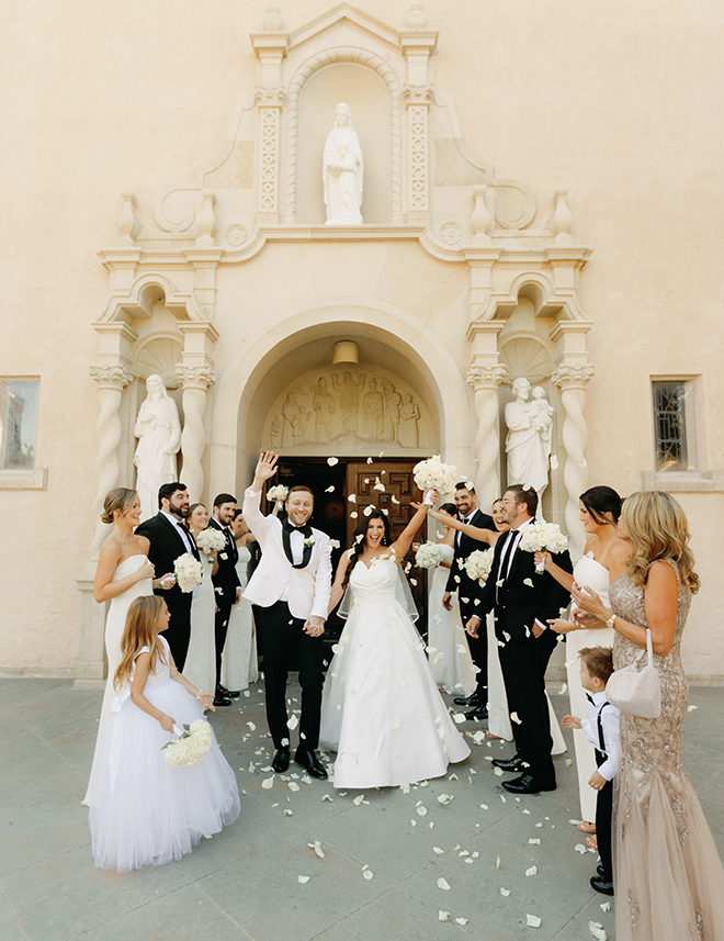 The bride and groom cheering while coming out of the church while her guests throw white rose petals. 