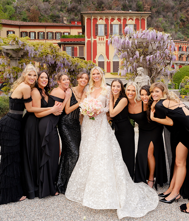 Eight bridesmaids wearing black dresses hugging the bride in the center. 