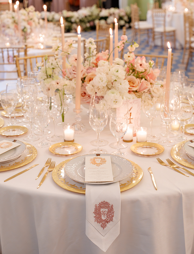 A round reception table with white linens, gold plates and flatware, pink candles and delicate floral centerpieces. 