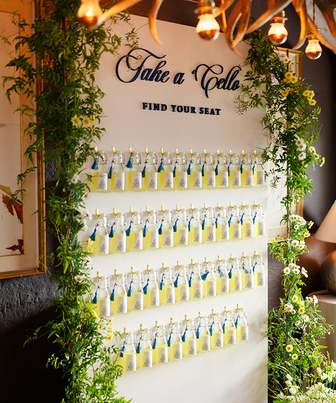 A wall of mini limoncello bottles on a wall that says "Take a Cello. Find Your Seat" in blue lettering. 