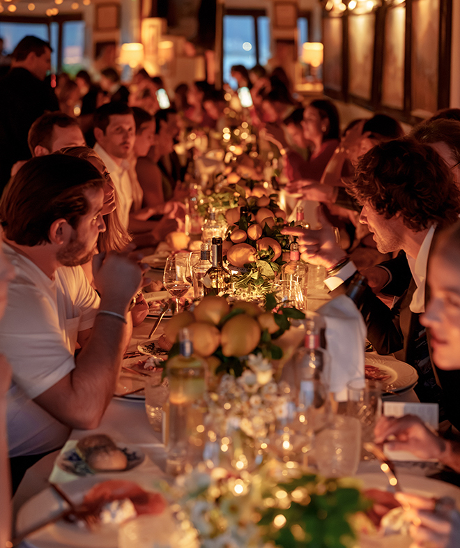 Guests dining at a long table with lemon centerpieces and warm lighting. 