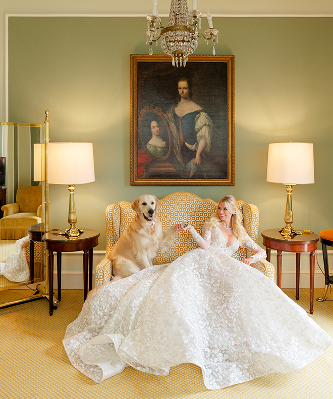 A bride sitting on a couch in a long sleeve lace wedding gown holding a golden retriever's paw. 