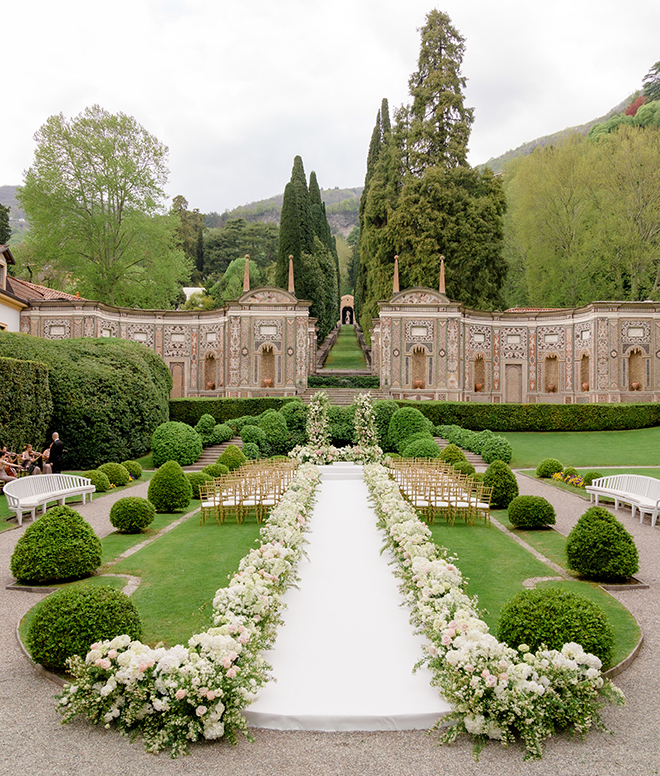 An aisle lined with white florals leading to an altar with a grand floral arrangement overlooking Lake Como. 