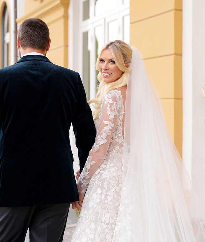 The bride holding the groom's hand looking back, wearing a long veil and a lace long-sleeve wedding gown. 