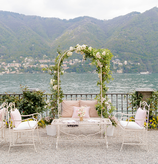 An archway decorated with greenery and white and pink furniture with a view of Lake Como for cocktail hour. 