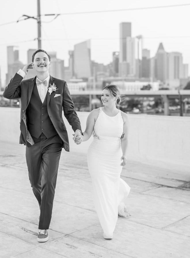 The bride and groom hold hands on the rooftop of their wedding venue overlooking the Houston skyline.