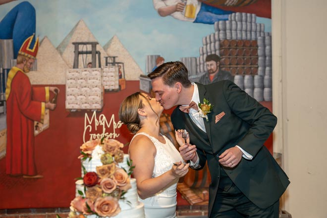 The bride and groom share a kiss beside their white wedding cake detailed in terracotta and tan flowers/