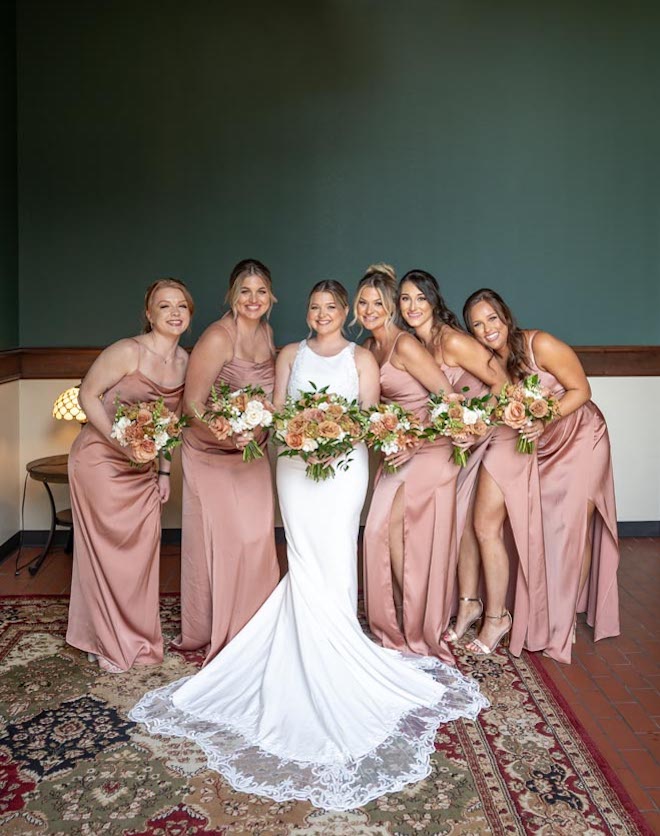 The bride and her bridesmaids hold wedding bouquets with terracotta, tan, gold, white & sage green flowers.