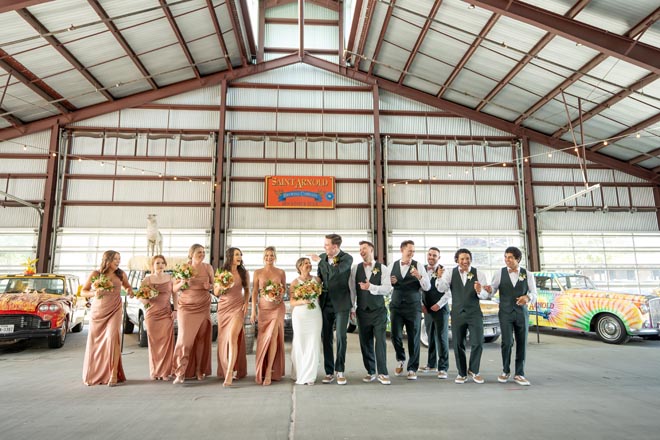 The couple and their wedding party walk in front of colorful vintage cars at their Houston wedding venue.