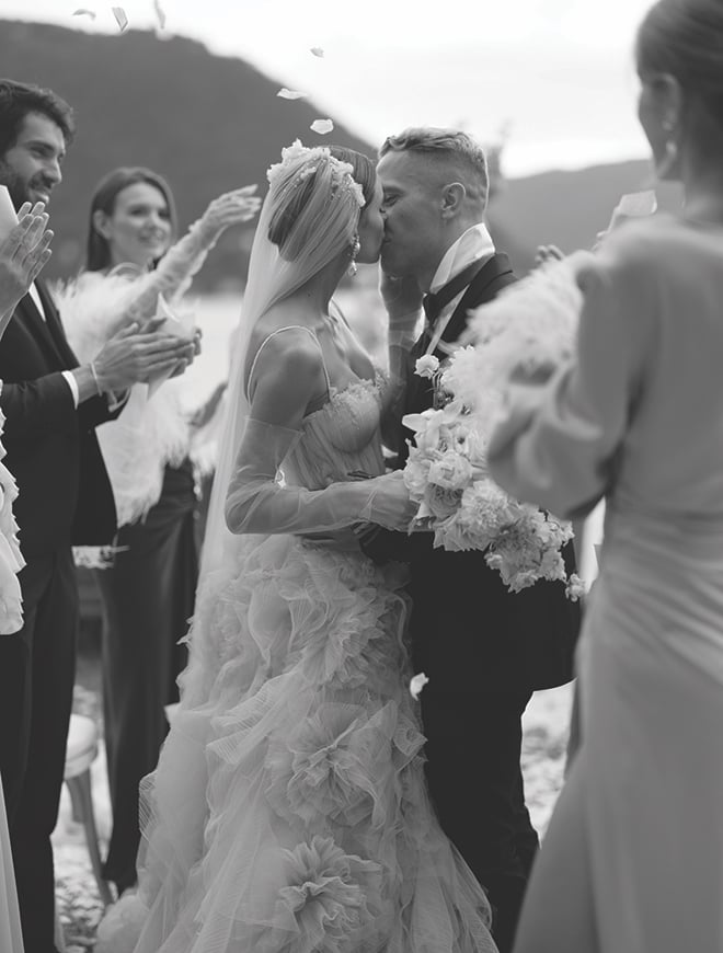 The bride and groom kissing while their guests cheer and throw flower petals. 